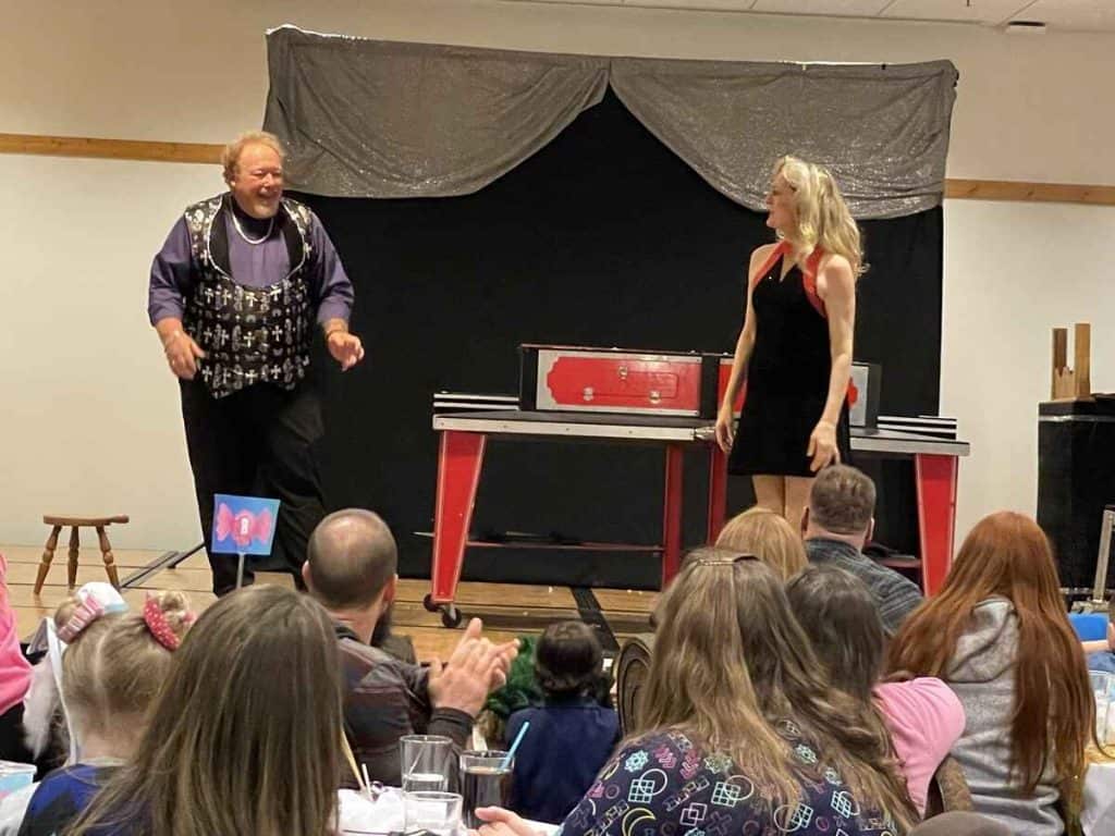 John Measner Magic Show | Why Hiring a Local Chicago Magician Makes Your Event Instantly More Memorable Magician John Measner in a purple vest and his assistant in a black dress smiling during a live magic show performance with an excited audience in the background.