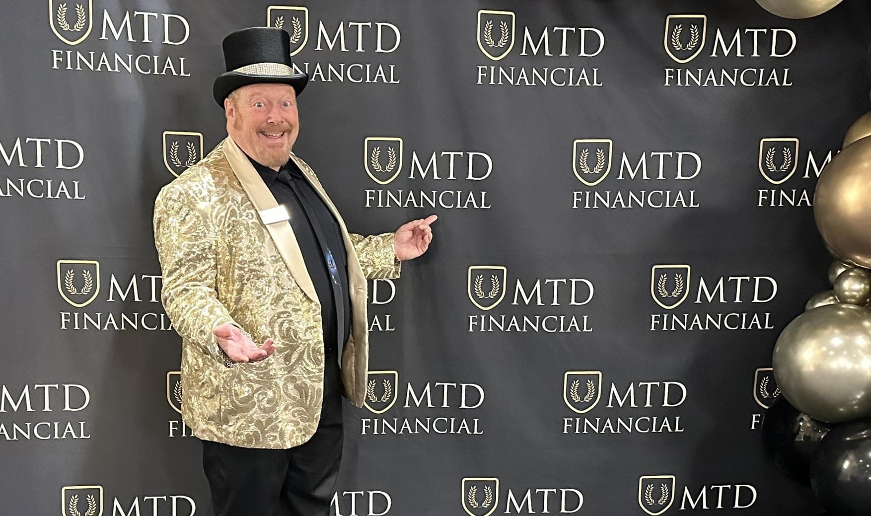 Corporate magician John Measner in a gold sequined jacket and top hat posing in front of an MTD Financial branded step-and-repeat backdrop