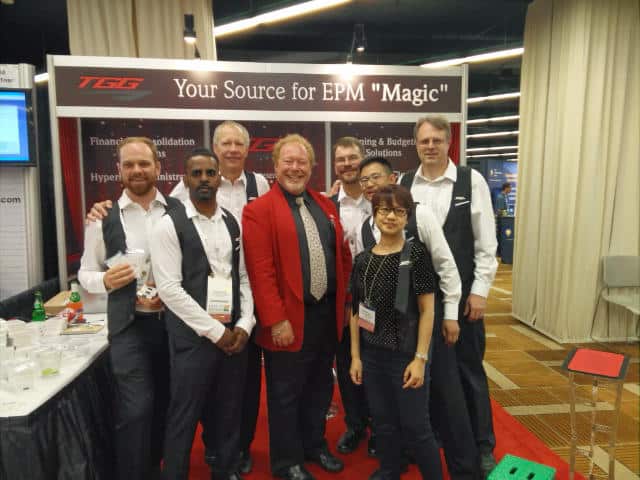 John Measner with the Client Booth Team at a Tradeshow Tradeshow magician John Measner, wearing his signature red jacket, poses with the booth team in front of a client display banner at a convention event.