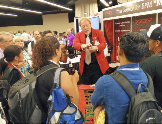 Magician Entertaining Crowd at Trade Show Booth A magician in a red jacket performs a close-up trick for a small crowd gathered around a booth at a busy trade show, with attendees watching closely and engaging with the performance.